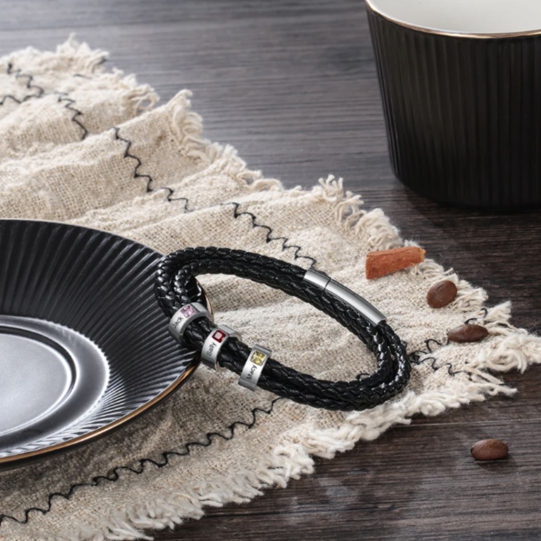 La Pulsera Piedras de vida - A mi Padre, una pulsera de cuero trenzado negro con detalles plateados, descansa sobre una tela beige con flecos cerca de granos de café y canela, lo que la convierte en un regalo ideal Día del Padre.