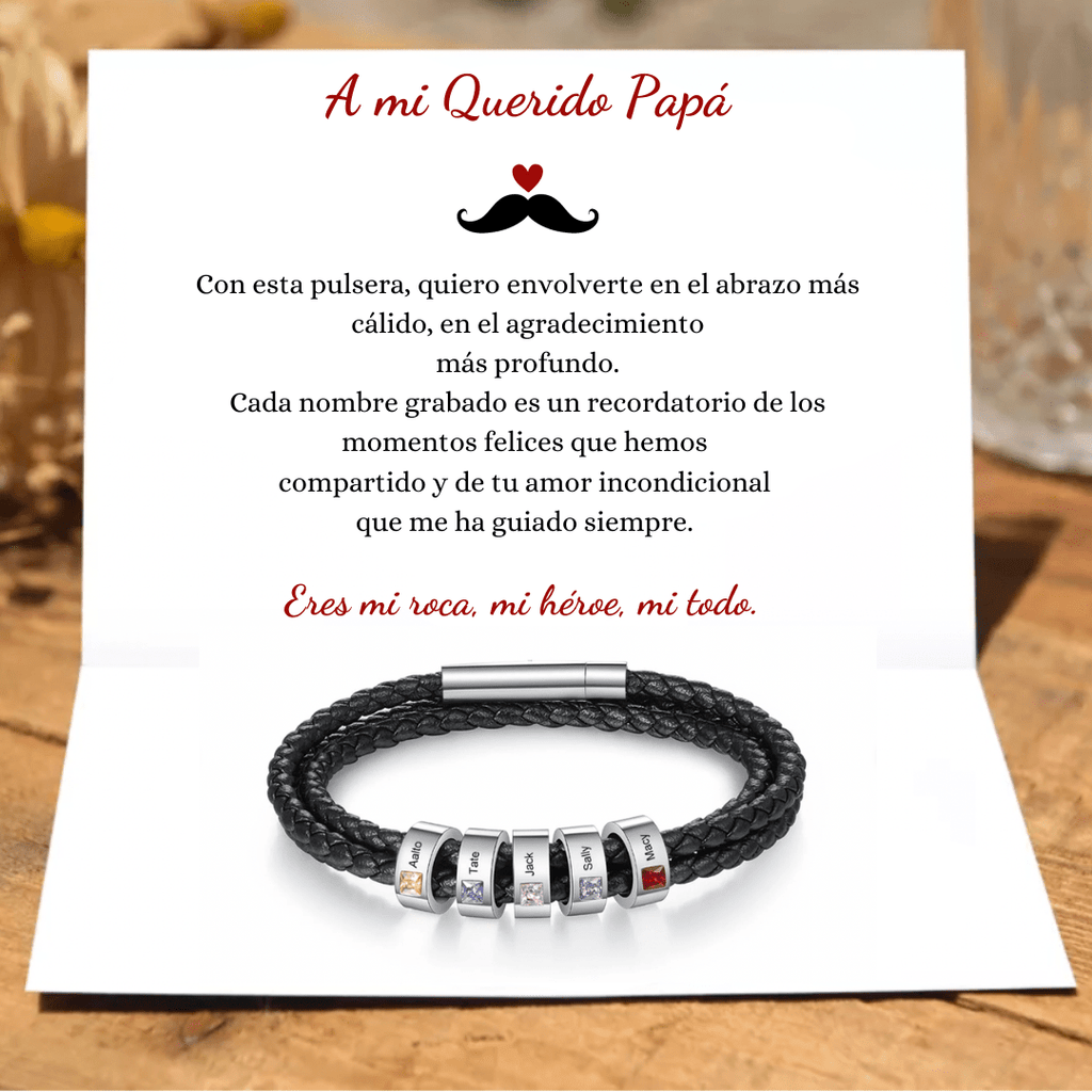 Una tarjeta con mensaje para papá y la Pulsera Piedras de vida - A mi Padre, negra trenzada con cuentas grabadas y detalles de colores. Al fondo, flores amarillas desenfocadas realzan este regalo especial del Día del Padre.