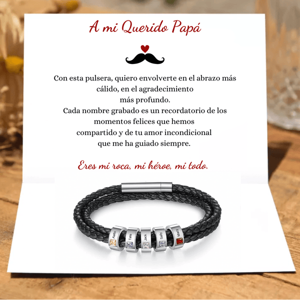Una tarjeta con mensaje para papá y la Pulsera Piedras de vida - A mi Padre, negra trenzada con cuentas grabadas y detalles de colores. Al fondo, flores amarillas desenfocadas realzan este regalo especial del Día del Padre.