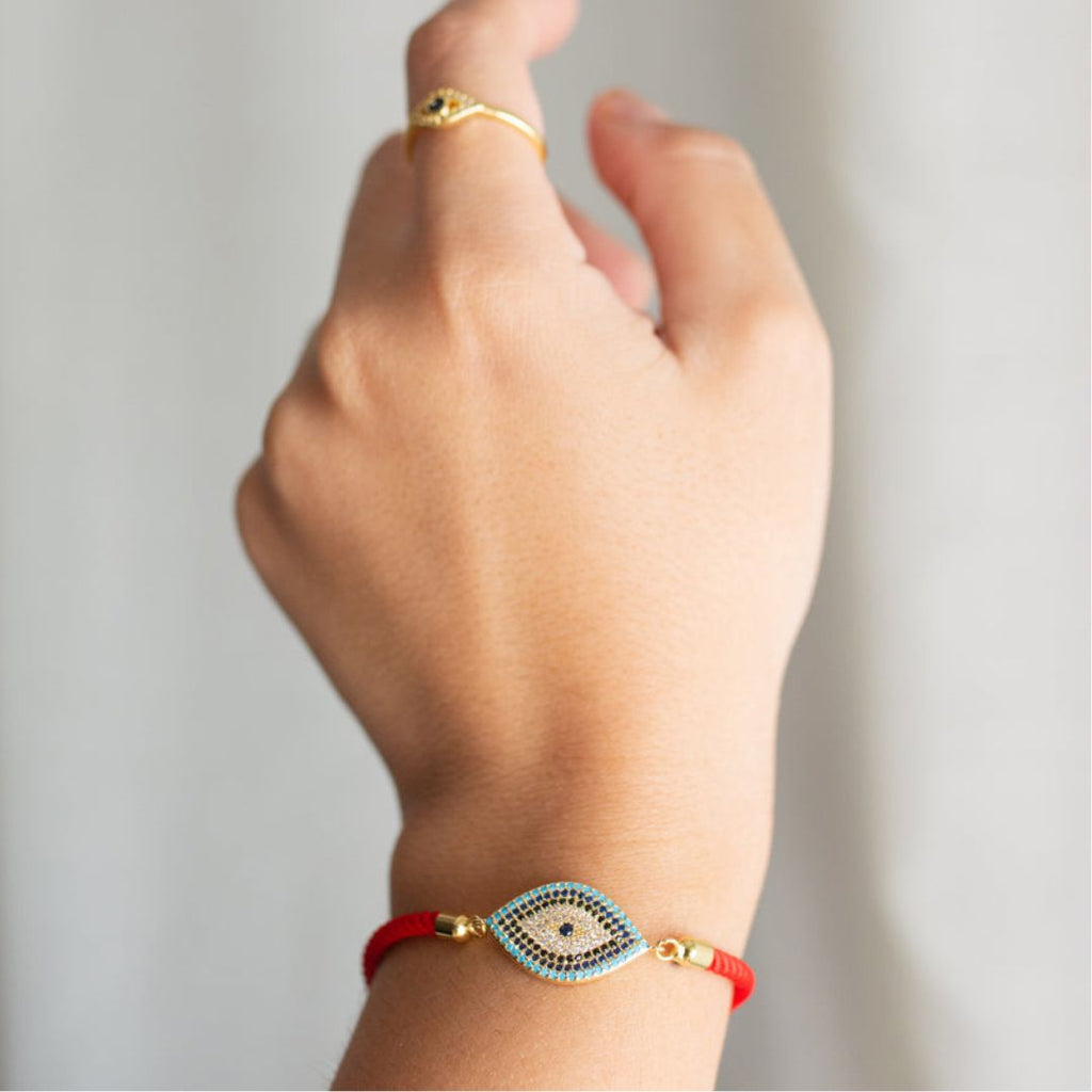 Una mano con un anillo de oro y la Pulsera protectora de ojo turco -una pulsera roja con un amuleto de ojo con piedras azules y blancas- destaca sobre un fondo suavemente difuminado.