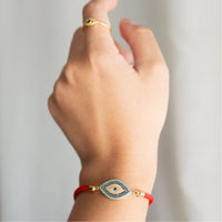 Una mano con un anillo de oro y la Pulsera protectora de ojo turco -una pulsera roja con un amuleto de ojo con piedras azules y blancas- destaca sobre un fondo suavemente difuminado.