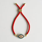 La Pulsera protectora de ojo turco es una pulsera roja con detalles dorados y un colgante en forma de ojo, con piedras azules, blancas y doradas: una elegante pulsera protectora que se muestra sobre un fondo blanco.