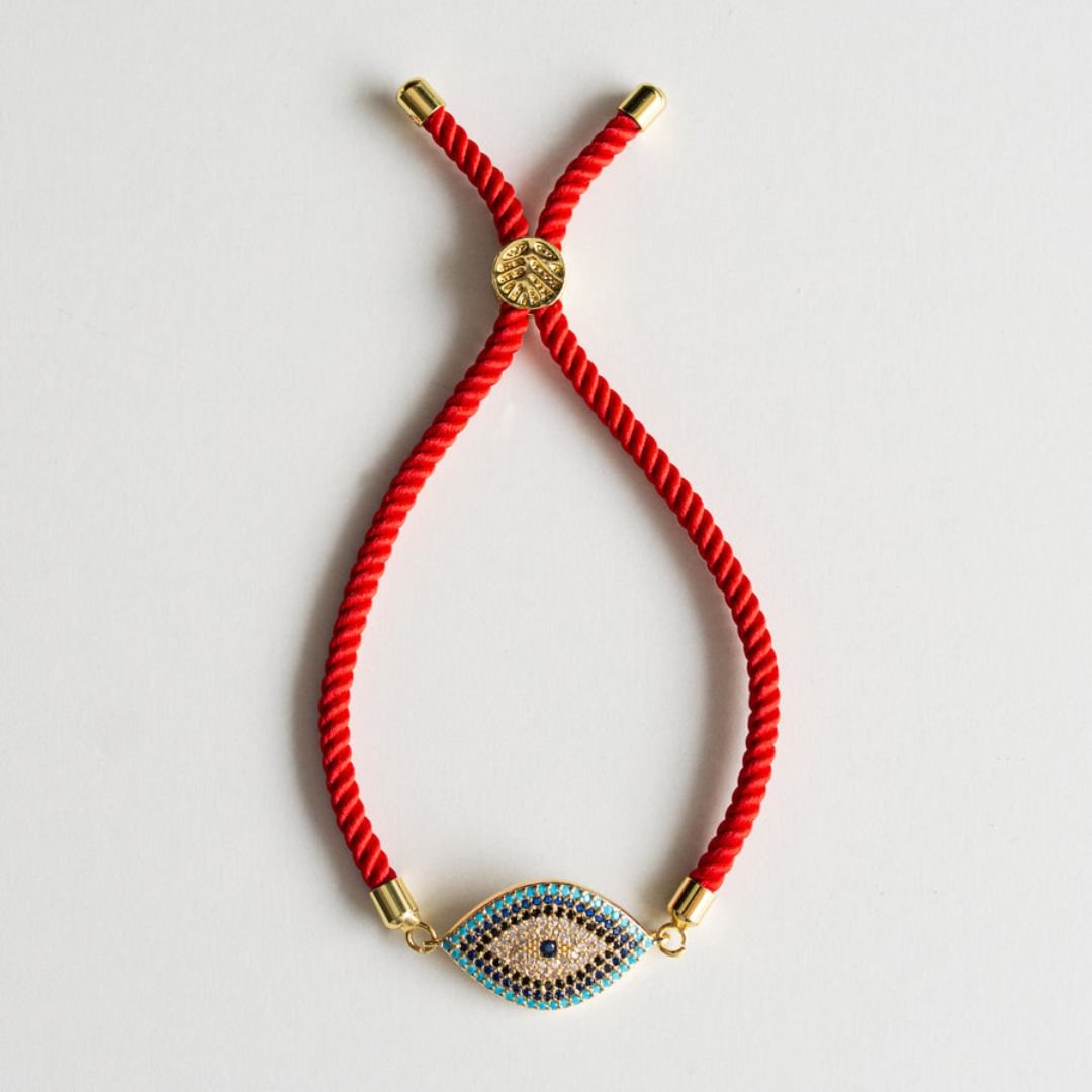La Pulsera protectora de ojo turco es una pulsera roja con detalles dorados y un colgante en forma de ojo, con piedras azules, blancas y doradas: una elegante pulsera protectora que se muestra sobre un fondo blanco.