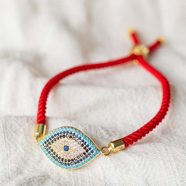 La Pulsera protectora de ojo turco presenta una banda roja con un amuleto de mal de ojo dorado, azul y blanco adornado con brillantes strass, sobre una suave tela blanca: un elegante accesorio de estilo y protección.