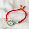La Pulsera protectora de ojo turco presenta una banda roja con un amuleto de mal de ojo dorado, azul y blanco adornado con brillantes strass, sobre una suave tela blanca: un elegante accesorio de estilo y protección.