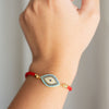 Una mano lleva la Pulsera protectora de ojo turco, una pulsera roja con un amuleto de mal de ojo dorado y azul sobre un fondo claro y liso. Se cree que esta elegante pieza trae buena suerte.