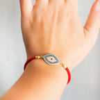 Una mano luce la Pulsera protectora de ojo turco, una pulsera de cordón rojo con un charm en forma de ojo azul, negro y dorado. El fondo suavemente difuminado resalta este elegante accesorio.