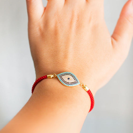 Una mano luce la Pulsera protectora de ojo turco, una pulsera de cordón rojo con un charm en forma de ojo azul, negro y dorado. El fondo suavemente difuminado resalta este elegante accesorio.