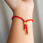 Primer plano de una muñeca que luce la Pulsera protectora de ojo turco, con cordón trenzado rojo, puntas metálicas doradas y una cuenta central redonda dorada. El fondo suavemente difuminado resalta este elegante accesorio.