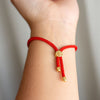 Primer plano de una muñeca que luce la Pulsera protectora de ojo turco, con cordón trenzado rojo, puntas metálicas doradas y una cuenta central redonda dorada. El fondo suavemente difuminado resalta este elegante accesorio.