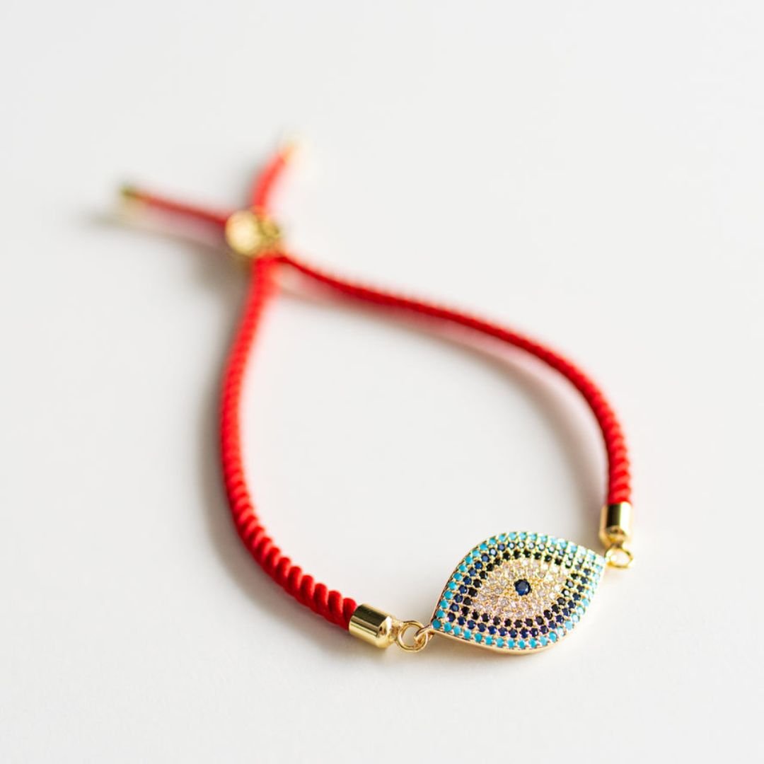 Pulsera protectora de ojo turco roja ajustable con amuleto de mal de ojo dorado y azul acentuado con pequeñas piedras preciosas, sobre fondo blanco.