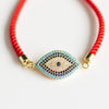 Pulsera protectora de ojo turco en color rojo con dije dorado y azul, adornada con piedras brillantes, sobre fondo blanco.