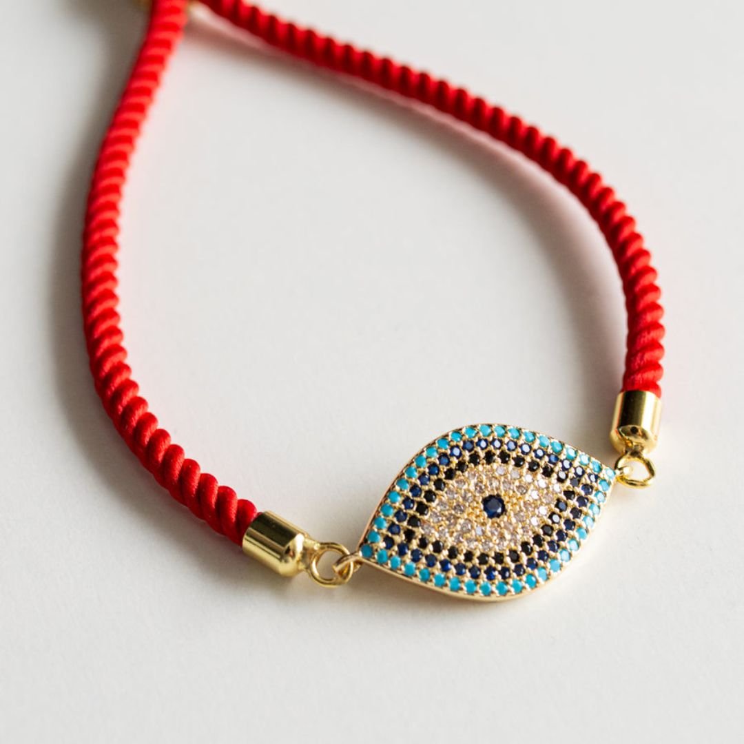La Pulsera protectora de ojo turco es una pulsera roja con dije dorado y turquesa de ojo turco, decorada con pequeñas piedras azules y negras para brindar estilo y protección, exhibida sobre una superficie blanca.