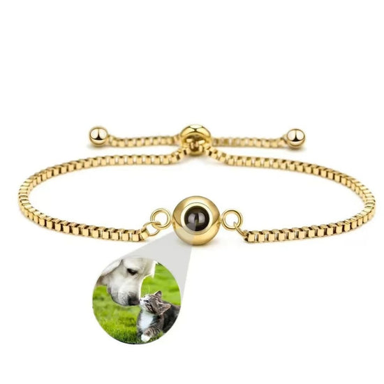 La Pulsera Recuerdo eterno en dorado tiene un colgante redondo con una pequeña foto de un perro y un gato tocándose las narices sobre hierba verde, ideal como regalo personalizado para los amantes de los animales.