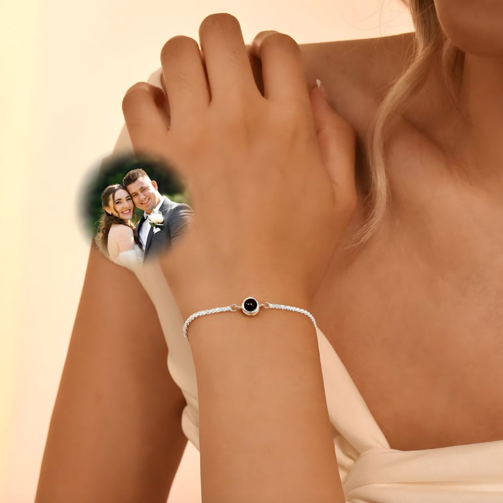 Una mujer que lleva la elegante Pulsera Recuerdo eterno descansa la mano sobre su hombro. Una superposición difuminada de una pareja de novios sonrientes sugiere que esta pulsera es un significativo regalo personalizado.