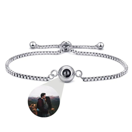 La Pulsera Recuerdo eterno en color plateado tiene un dije central redondo que proyecta una imagen personalizada, como una pareja abrazada al aire libre-un regalo personalizado memorable.