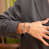Una persona con americana gris y puños abotonados muestra una mano adornada con anillos de plata y la Pulsera Riviera, con varias pulseras, sobre un fondo borroso de hojas verdes.