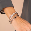 Primer plano de una muñeca adornada con tres pulseras de plata, entre ellas la Pulsera Riviera con pequeñas tachuelas, además de un diseño con textura y otro con acabado liso. La portadora va vestida con un top gris de manga larga.