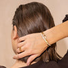 Una persona de pelo castaño corto, vista de espaldas, se toca la nuca, luciendo anillos de oro y dos pulseras -una de ellas es la Pulsera Riviera- sobre un fondo neutro.