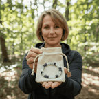 En un bosque iluminado por el sol, una mujer muestra una pequeña bolsa con cordón que contiene Pulsera Serena - Alivio natural para la menopausia, una pulsera de cuentas con piedras naturales moradas y blancas.