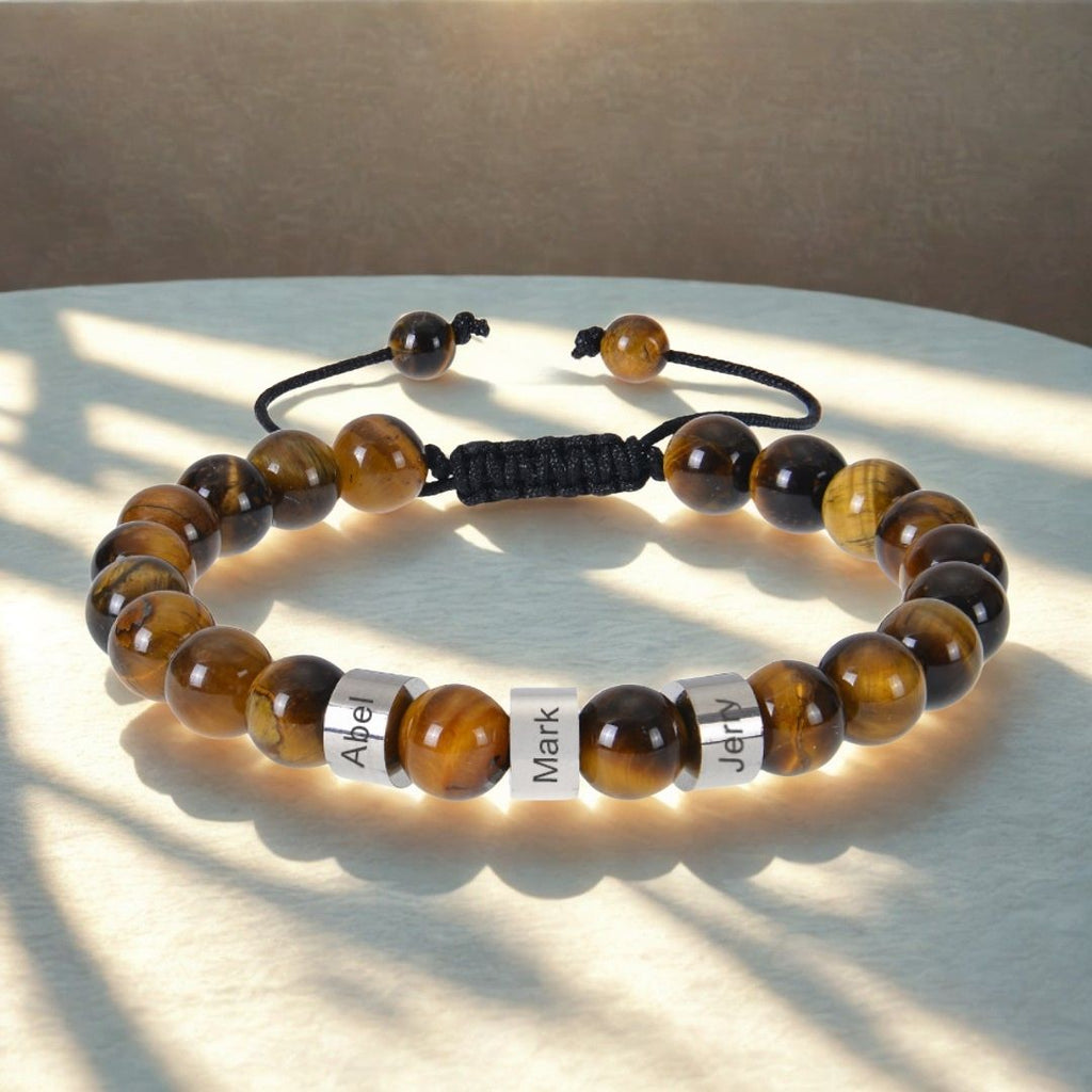 A Pulsera tejida personalizada con nombres, hecha con cuentas de ojo de tigre marrón y dorado pulido y tres abalorios plateados grabados con Abel, Mark y Jerry, es una pulsera especial para papá exhibida bajo la luz del sol.