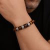 Primer plano de una muñeca que lleva una Pulsera tejida personalizada con nombres con cuentas marrones y plateadas, algunas grabadas. La persona, que tiene vello facial, apoya la barbilla en el brazo para resaltar esta pulsera personalizada para papá.