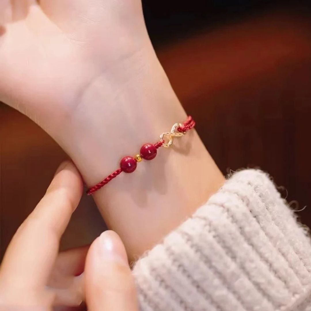 Primer plano de una muñeca que lleva una Pulsera de amor infinito - Para Él y para Ella, con un diseño trenzado rojo con dos cuentas rojas y detalles dorados, junto a una mano que la ajusta. La persona lleva un jersey de canalé beige claro.