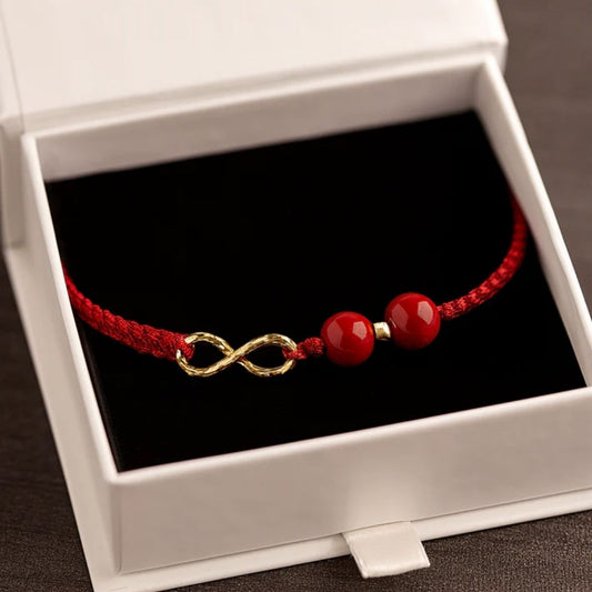 Pulseras de amor infinito - Para Él y para Ella: Una pulsera roja trenzada con dos cuentas rojas brillantes y un charm dorado infinito, presentada en un joyero blanco abierto con interior negro.