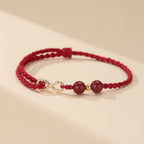 Las Pulseras de amor infinito - Para Él y para Ella presentan un diseño trenzado rojo con dos cuentas redondas rojas, una pequeña cuenta dorada y un colgante dorado con el símbolo del infinito, sobre un fondo beige claro.