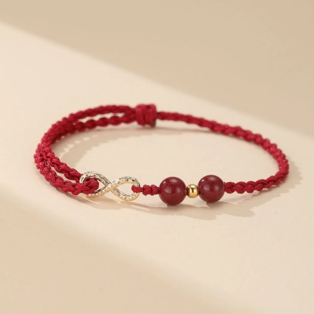 Las Pulseras de amor infinito - Para Él y para Ella presentan un diseño trenzado rojo con dos cuentas redondas rojas, una pequeña cuenta dorada y un colgante dorado con el símbolo del infinito, sobre un fondo beige claro.