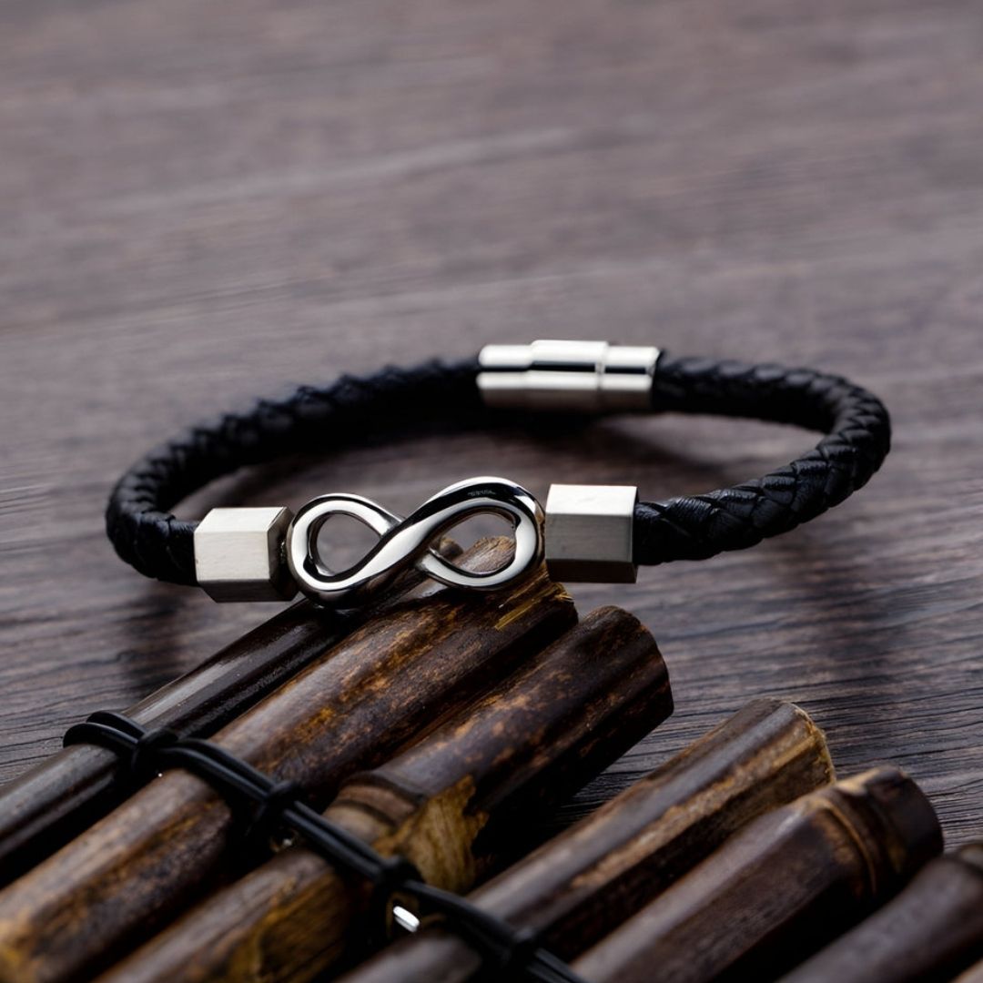 Pulseras de amor infinito - Para Él y para Ella: una pulsera de cuero negro trenzado con símbolo infinito plateado y cierre metálico, sobre madera oscura junto a varas de bambú.