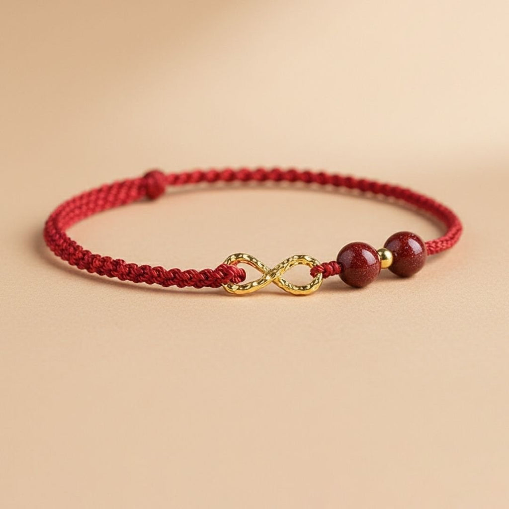 Pulseras de amor infinito - Para Él y para Ella: pulsera de cordón trenzado rojo con una pieza central dorada del símbolo del infinito y dos cuentas redondas rojas, presentada sobre una superficie neutra de color beige.
