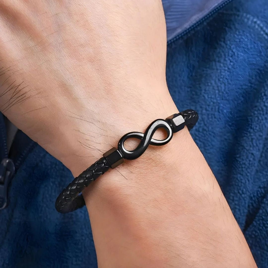Una mano lleva una Pulsera de amor infinito - Para Él y para Ella, con una banda trenzada negra con un símbolo metálico del infinito, sobre tela azul.