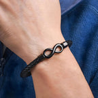 Una mano lleva una Pulsera de amor infinito - Para Él y para Ella, con una banda trenzada negra con un símbolo metálico del infinito, sobre tela azul.