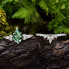 Sobre madera oscura, el Set de anillos piedra ágata verde resalta: uno de compromiso con piedra verde y diamantes blancos, y otro curvo con pequeños diamantes. Follaje natural enmarca este elegante conjunto de anillos.