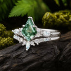 El set de anillos piedra ágata verde en color plateado destaca por su gema central en forma de cometa con vetas musgosas, rodeada de pequeñas piedras claras y presentada sobre una roca oscura con follaje verde al fondo.