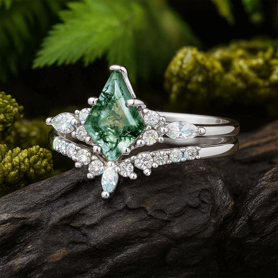 El set de anillos piedra ágata verde en color plateado destaca por su gema central en forma de cometa con vetas musgosas, rodeada de pequeñas piedras claras y presentada sobre una roca oscura con follaje verde al fondo.