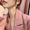 Primer plano de una mujer vestida con un blazer rosa rubor y el Set de aretes brillantes, con la barbilla apoyada en la mano mientras una suave luz natural resalta su rostro y su pelo.
