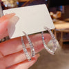 Una mano sostiene una tarjeta blanca en la que aparece el Set de aretes brillantes, unos pendientes de aro plateados con brillantes transparentes. El fondo suavemente difuminado acentúa el brillo y la elegancia de estos pendientes brillantes.