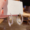 Una mano sostiene una tarjeta blanca que muestra un Set de aretes brillantes en color plateado, adornado con brillantes strass, sobre un fondo suavemente difuminado.