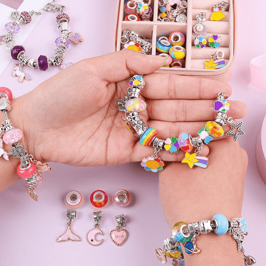 Un niño sostiene una pulsera colorida hecha con el set de joyería para crear pulseras y collares + accesorios, mientras más pulseras, dijes y una caja rosa se exhiben sobre un fondo pastel; ideal para fomentar habilidades motrices.