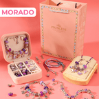 El Set de joyería para crear pulseras y collares + accesorios se muestra con pulseras, dijes y collares en tonos morados, junto a una bolsita de regalo rosa y caja joyero sobre fondo rosado. "MORADO" resalta en letras blancas.