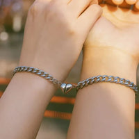 Dos manos lucen el Set de pulseras con corazón magnético, ambas cadenas plateadas con dijes de corazón, mostradas juntas contra un fondo exterior desenfocado-el regalo perfecto para parejas.