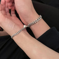 Dos manos entrelazadas lucen el set de pulseras con corazón magnético-cadenas plateadas con dijes de corazón-sobre un fondo oscuro, ideal como regalo para parejas.