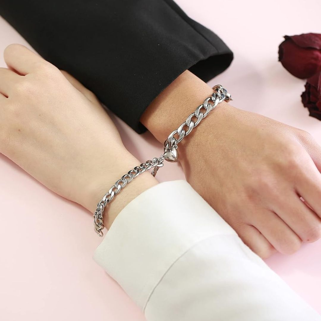 Dos manos, una en negro y otra en blanco, llevan el Set de pulseras con corazón magnético enlazadas por un charm de corazón, descansando sobre una superficie rosa claro junto a rosas rojas secas-el regalo ideal para parejas.