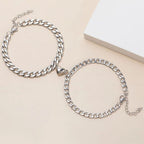El Set de pulseras con corazón magnético, color plateado, es ideal como regalo para parejas; incluye dos pulseras ajustables mostradas sobre una superficie beige, una con charm de corazón en el centro.