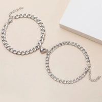 El Set de pulseras con corazón magnético, color plateado, es ideal como regalo para parejas; incluye dos pulseras ajustables mostradas sobre una superficie beige, una con charm de corazón en el centro.
