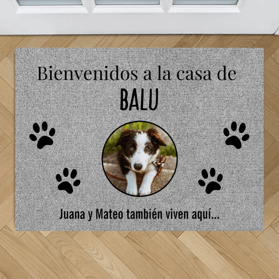 Tapete personalizado con fotos de mascotas: tapete color gris con huellas negras y foto de perro marrón y blanco en el centro. Texto: "Bienvenidos a la casa de BALU. Juana y Mateo también viven aquí...". Ideal para mascotas.